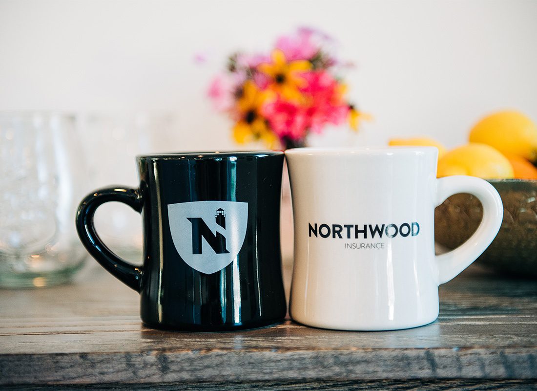 About Our Agency - Closeup View of Two Different Colored Mugs with the Northwood Insurance Office Decal Sitting on a Wooden Table Inside the Office