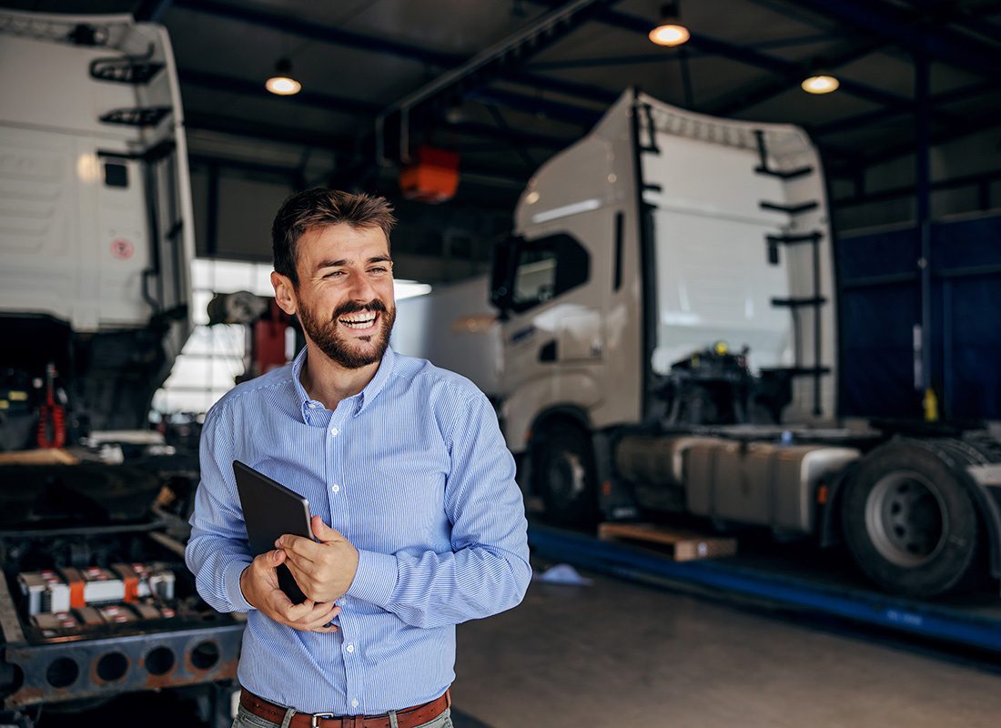 Business Insurance - Closeup Portrait of a Smilng Young Businessman Standing in a Garage with Two Semi Trucks While Holding a Tablet in his Hands