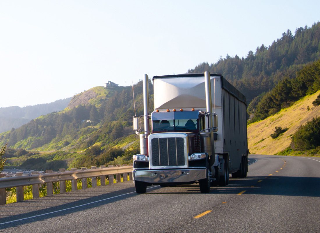 Insurance by Industry - Closeup View of a Modern Semi Truck with a Trailer Driving on a Scenic Empty Highway by the Coast with Views of Mountains Covered with Green Trees
