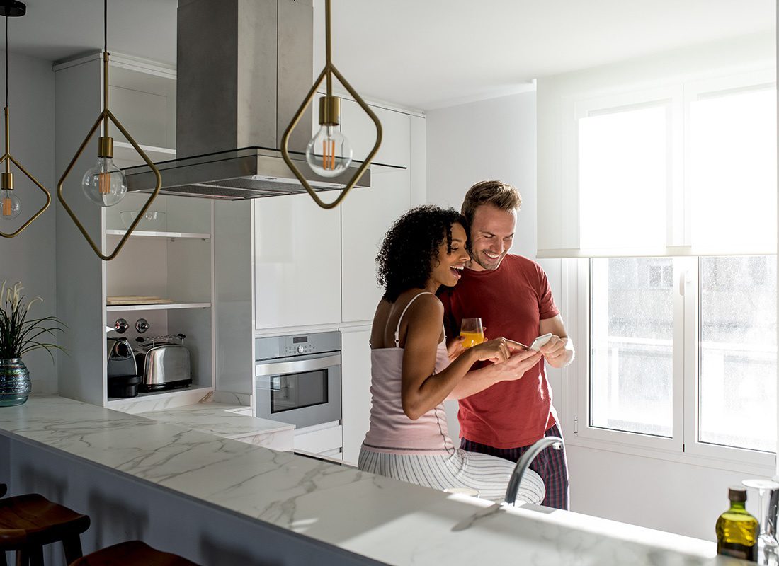 Personal Insurance - Portrait of a Cheerful Young Married Couple Using a Phone While Spending Time Together in their Modern Kitchen in the Morning